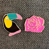 Toekan Emaille Pin Licht Roze Snavel samen met een roze roos pin op een grijze ondergrond