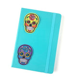 Twee strijk patches van een rijkversierde gele en paarse sugar skull. Met geborduurde versierde  accenten op de voorzijde van een blauw groen notitieboekje